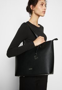 Schwarze Leder-Tote mit strukturiertem Design, silbernen Hardware-Details und glatter Textur. Mit einem Logo-Detail auf der Vorderseite.