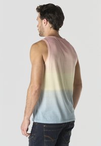 Hombre visto desde atrás que lleva una camiseta sin mangas con un degradado en tonos rosa, amarillo y azul, combinada con vaqueros azul oscuro frente a un fondo liso.