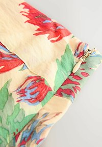 Blumenmusterstoff in Creme, mit roten und blauen Blumen sowie grünen Akzenten. Enthält einen verdeckten Reißverschluss mit einem beigen Ziehband.