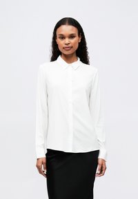 Chemise blanche à manches longues avec un col pointu, fermeture à boutons sur le devant et tissu lisse. Associe à une jupe noire.