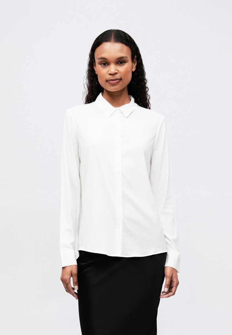 Chemise blanche à manches longues avec un col pointu, fermeture à boutons sur le devant et tissu lisse. Associe à une jupe noire.