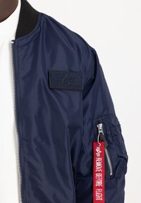 Blå nylon bomberjakke med ribbet krave, et logopatch på brystet og en lynlås lomme med et rødt "REMOVE BEFORE FLIGHT" mærkat.