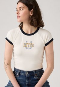 Hvid tætsiddende T-shirt med marineblå kant og korte ærmer. Har et centralt broderet grafik af en hestesko og "LEVI'S" i gul.