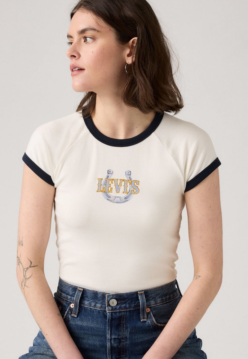 Hvid tætsiddende T-shirt med marineblå kant og korte ærmer. Har et centralt broderet grafik af en hestesko og "LEVI'S" i gul.