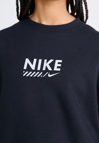 Svart sweatshirt med rund hals, som har en framträdande vit "NIKE" logotyp med ett randigt mönster under och en liten swoosh-accent.