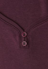 Bordeaux strikstof med V-hals-design og to lilla knapper. Glat tekstur med subtile syninger omkring halsudskæringen.