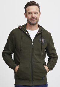 Maslinasto zelena zip-up dukserica s podstavljenom kapuljačom, logotipom, prednjim džepovima i rebrastim manžetama; izrađena od mekog, udobnog materijala.