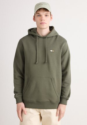 Giovane uomo che indossa una felpa verde oliva con tasca frontale e pantaloni beige, in piedi su uno sfondo chiaro e semplice.