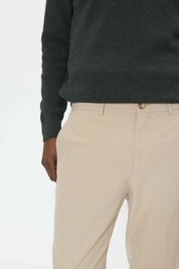 Osoba ubrana w beżowe spodnie i ciemnoszary, dziany sweter z długim rękawem, stojąca z jedną ręką swobodnie przy boku.