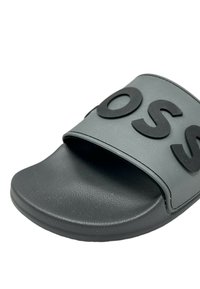 Sandale en caoutchouc grise avec un lit de pied texturé, une large sangle ornée de lettres noires en relief et un design de semelle lisse et arrondie.