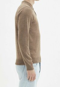 Pull en tricot marron à manches longues, col et ourlet côtelé. Porté avec un jean bleu clair. Texture lisse, coupe ajustée.