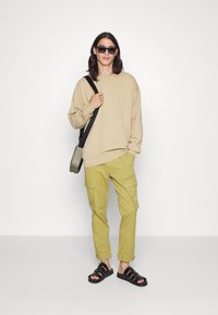 Sweatshirt oversize beige, calças cargo verde claro com bolsos, e sandálias pretas. Os acessórios incluem óculos de sol pretos e uma mala de ombro preta.