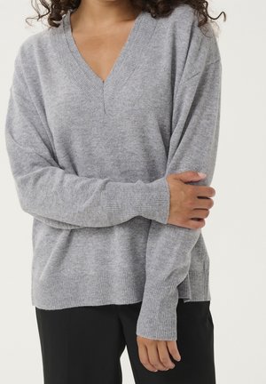 Personne portant un pull gris ample à col en V avec poignets et ourlet côtelés, un bras croisé tenant la manche opposée.