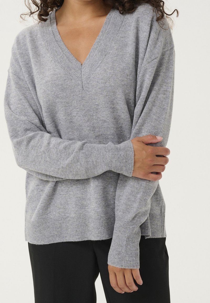 Personne portant un pull gris ample à col en V avec poignets et ourlet côtelés, un bras croisé tenant la manche opposée.