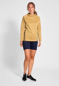 Gul huvtröja med svarta dragkedjedetaljer på sidan, kombinerad med mörkblå shorts. Modellen har på sig svarta sportskor. Minimalistisk design.