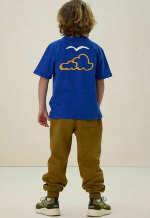 Kind met golvend haar staat met de rug naar de camera en draagt een blauw T-shirt met wolk- en vogelafbeelding, een bruine broek en groene sneakers tegen een effen achtergrond.