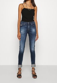 Jeans de denim ajustados en azul oscuro con un lavado deslavado, que presentan detalles desgastados y dobladillos enrollados, combinados con una blusa negra de tirantes finos.
