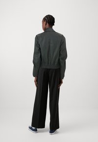 Tamsiai žalia bomberio tipo striukė su prigludusia liemeniu, ilgomis rankovėmis ir apykakle, derinama su juodomis dryžuotomis plačiomis kelnėmis.