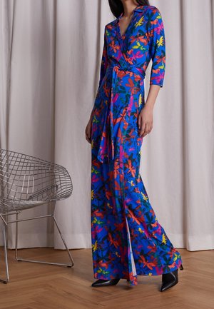 Woman wearing a blue floral wrap dress with three-quarter sleeves and black pointed-toe boots, standing near a silver wire chair.