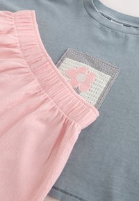 Pinkki kerätty hame, joka lepää harmaan paidan päällä, jossa on neliön muotoinen virkattu paikka, jossa keskellä on pinkki kukka.