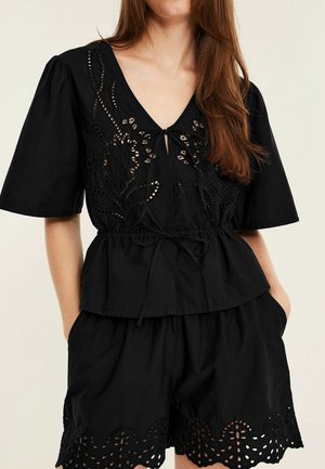 Vrouw draagt een zwart setje met een top met korte mouwen en korte broek met bloemrijke cut-out borduursel en een gestrikt taille detail.