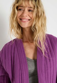 Gilet violet texturé avec des rayures verticales, ouvert à l'avant, porté sur un haut gris ajusté. Le tissu semble léger et respirant.