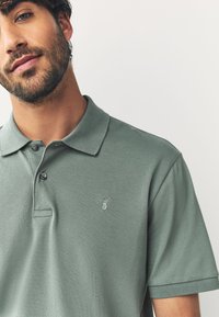 Pikétröja i dämpad grön bomull med knappslå och korta ärmar, med en broderad hjortlogotyp på bröstet. Texturerat tyg.