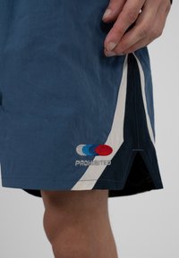 Nahaufnahme einer Hand neben blauen Shorts mit weißen und marineblauen Seitenpanelen sowie bestickten roten, blauen und weißen Kreisen mit dem Text "VERBOTEN".