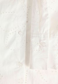 Λευκό βαμβακερό ύφασμα με κέντημα ματιών που απεικονίζει ανθικά μοτίβα και μικρά λευκά κουμπιά σε διπλωμένο τμήμα ενδύματος.