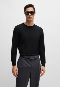 Crni pulover s okruglim izrezom od finog pleta, uparen sa sivim nabranim hlačama i crnim remenom. Model također nosi tamne naočale.