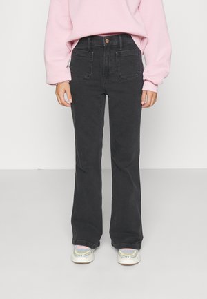 Person trägt schwarze hochtaillierte Jeans mit weitem Bein, rosa Sweatshirt und mehrfarbige Turnschuhe, steht vor einem schlichten Hintergrund.