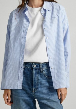 Femme portant une chemise à rayures bleu clair boutonnée par-dessus un t-shirt blanc et un jean bleu taille haute, debout devant un fond uni.