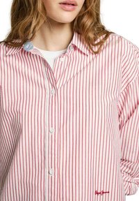Camicia button-up a righe rosse e bianche con colletto classico, dotata di un piccolo logo sul lato inferiore e un tessuto morbido e testurizzato.
