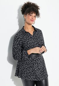 Schwarze Bluse mit einem grauen Blattmuster, mit Knopfleiste, großem Kragen und langen Ärmeln. Kombiniert mit einem schwarzen Ledergürtel.