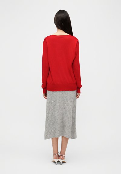 Κόκκινο πουλόβερ με μακριά μανίκια και άνετη εφαρμογή, συνδυασμένο με γκρι midi φούστα πλεκτή με σχέδιο κοτσίδα και άσπρα σανδάλια με τακούνι. Ομαλή υφή σε όλο το ύφασμα.