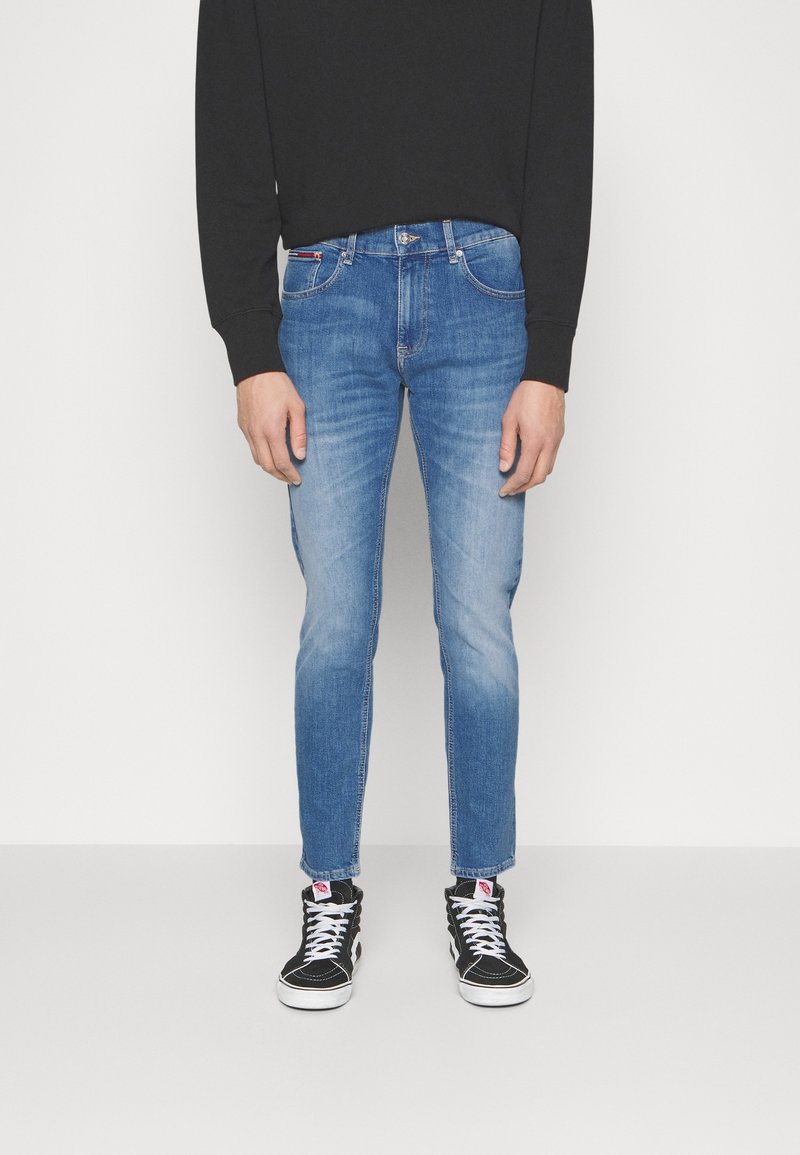 Blå denimjeans med en smal passform, blekta detaljer på låren och en klassisk femfickdesign, bärs med svarta högre sneakers.