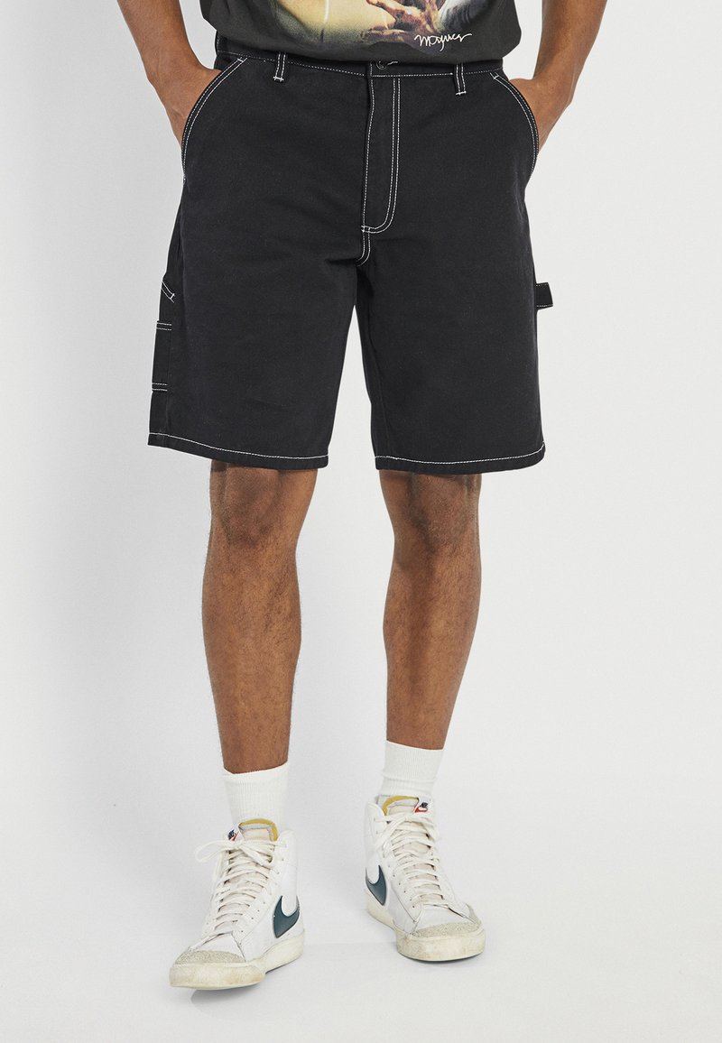 Mann trägt schwarze Shorts mit weißen Nähten, weiße High-Top-Sneakers und ein schwarzes Grafik-T-Shirt, steht mit den Händen in den Taschen.
