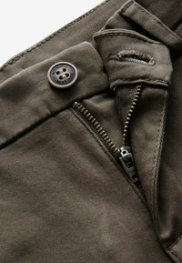 Pantalons vert olive foncé avec une fermeture à glissière en laiton, un bouton de fermeture et des détails de couture. Présente une étiquette avec le texte "AUTHENTIC NEXT DUDE".
