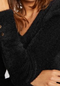 Schwarzer, flauschiger Strickpullover mit V-Ausschnitt und goldenen Knopfakzenten an den Ärmeln. Weiche Textur mit lockerer Passform.