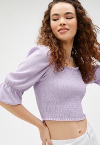 Lila cropped top met een smockeffect, pofmouwen en een gerimpelde zoom, gecombineerd met witte high-waisted broek.