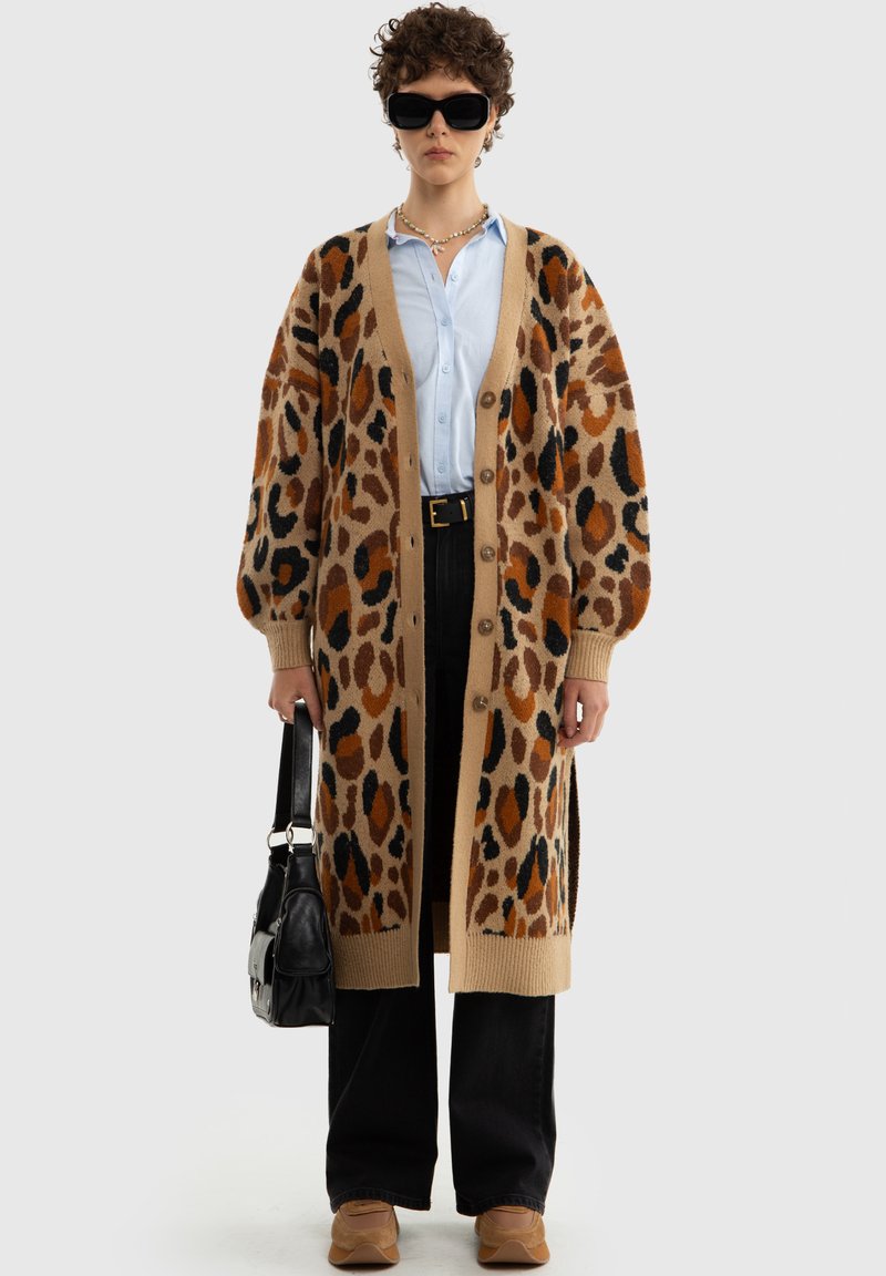 Pletený cardigan s leopardím vzorem v béžové, hnědé a černé barvě, s dlouhým, oversized fit, zapínáním na knoflíky a žebrovanými manžetami. Kombinovaný s světlomodrou košilí.