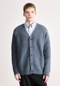 Γκρι πλεκτό cardigan με V λαιμόκοψη, έξι σκούρα κουμπιά και λάστιχο στους καρπούς, φτιαγμένο από μαλακό και αφράτο υλικό. Φοριέται πάνω από ένα ανοιχτόχρωμο ριγέ πουκάμισο.