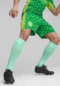 Zöld és sárga mintás focimez mentazöld zoknikkal. A zoknikon arany PUMA logó található; fekete focicipők textúrázott dizájnnal.