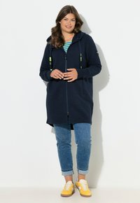 Námořnická mikina na zip s texturovanou tkaninou, vybavená kapucí, kapsami a neonovými akcenty na popruzích, doplněná světle modrými džíny a žlutými teniskami.