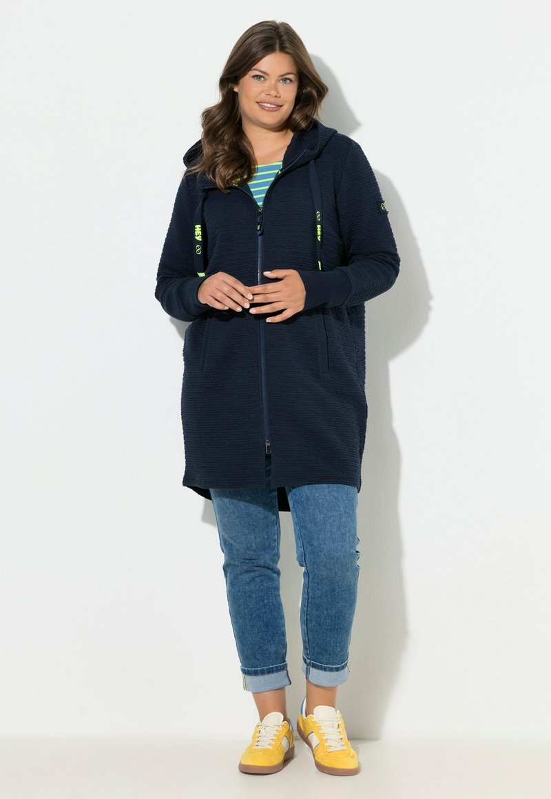 Námořnická mikina na zip s texturovanou tkaninou, vybavená kapucí, kapsami a neonovými akcenty na popruzích, doplněná světle modrými džíny a žlutými teniskami.