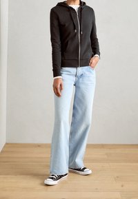 Schwarze Zip-Hoodie mit Fronttaschen, hellblaue weit geschnittene Jeans und schwarze Converse-Sneaker auf einem Holzboden vor einer hellen Wand.
