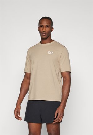 Beige cotton t-shirt with a crew neck, short sleeves, and EA7 logo in white on the chest. Paired with black shorts. Smooth texture.