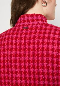 Tessuto a quadri rosso e rosa, caratterizzato da un colletto alto e un piccolo dettaglio di logo metallico sul retro. Finitura strutturata e motivo audace.