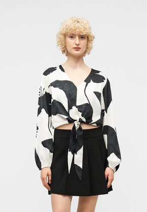 Persona bionda che indossa un crop top a maniche lunghe nero e bianco a fiori annodato e una gonna nera plissettata a vita alta su uno sfondo semplice.