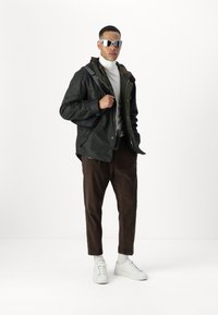 Dark green jacket with snap closures, layered over a gray sweater. Brown corduroy pants and white sneakers. Silver sunglasses and a black bag.